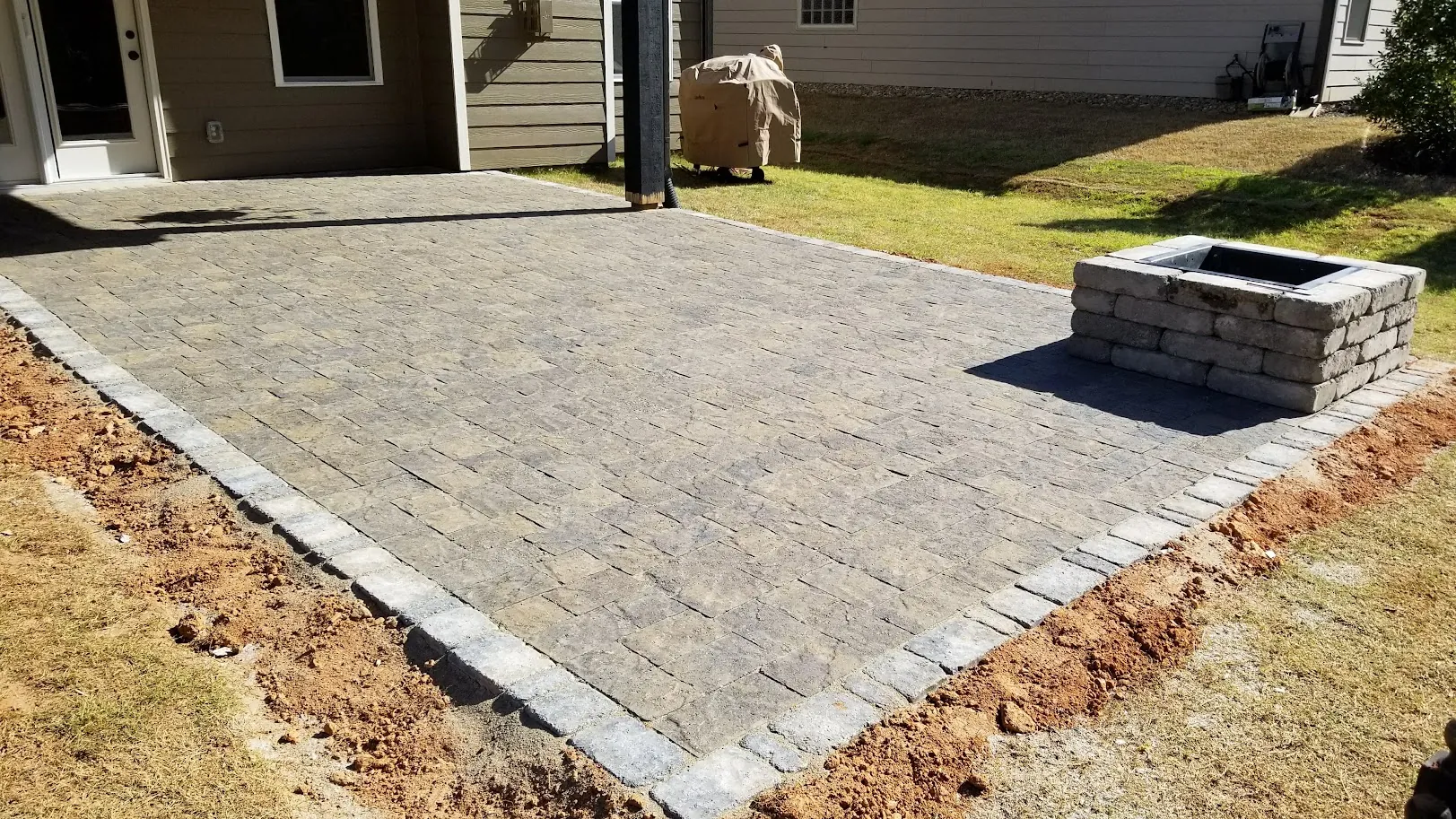Newly installed paver patio and fire pit, with disturbed soil still visible at the edges
