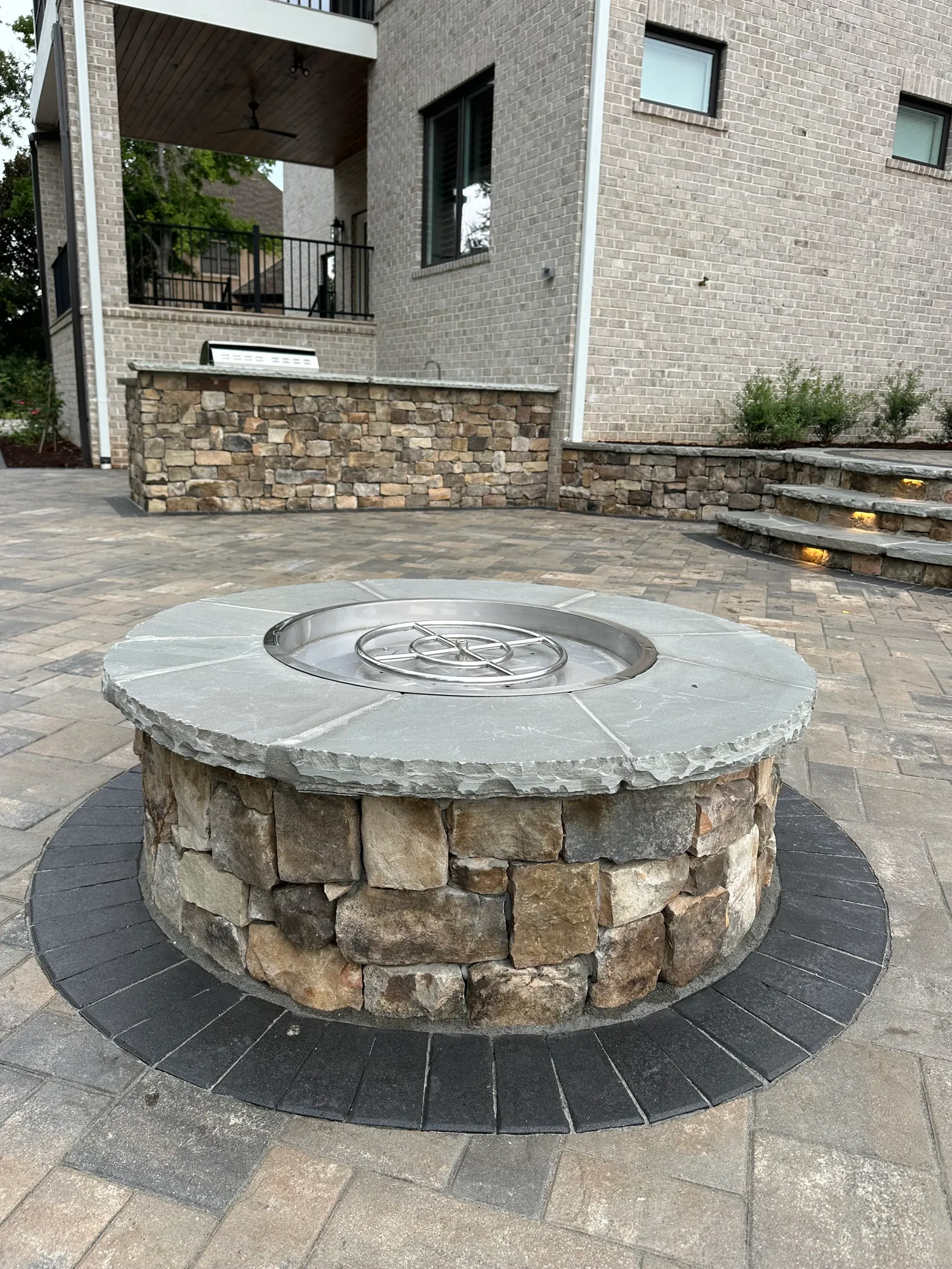 Round natural-stone fire pit with a gas insert on a paver patio, brick home with illuminated steps in the background