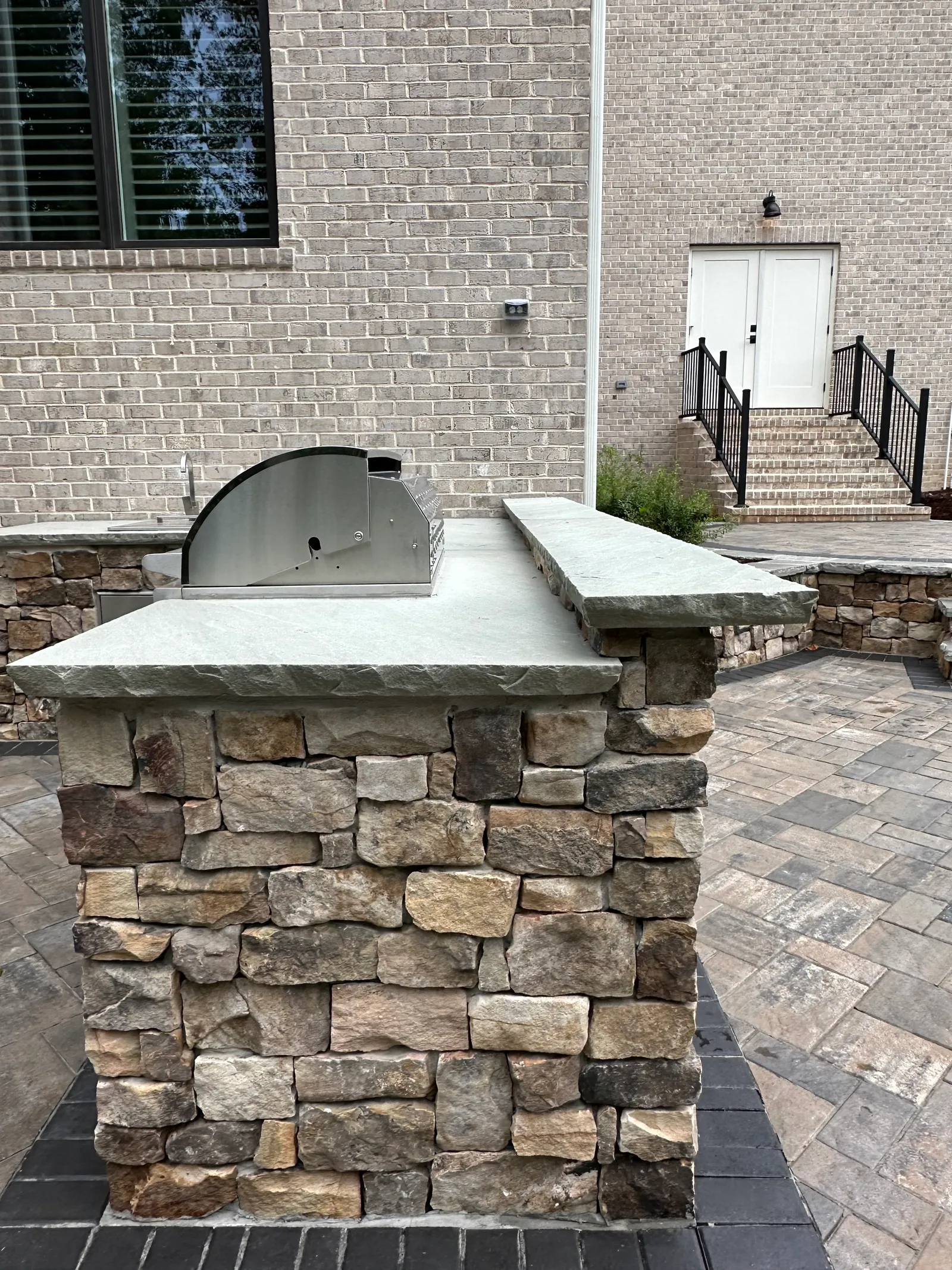Outdoor kitchen counter with a built-in gas burner on a paver patio, brick home with stone steps in the background