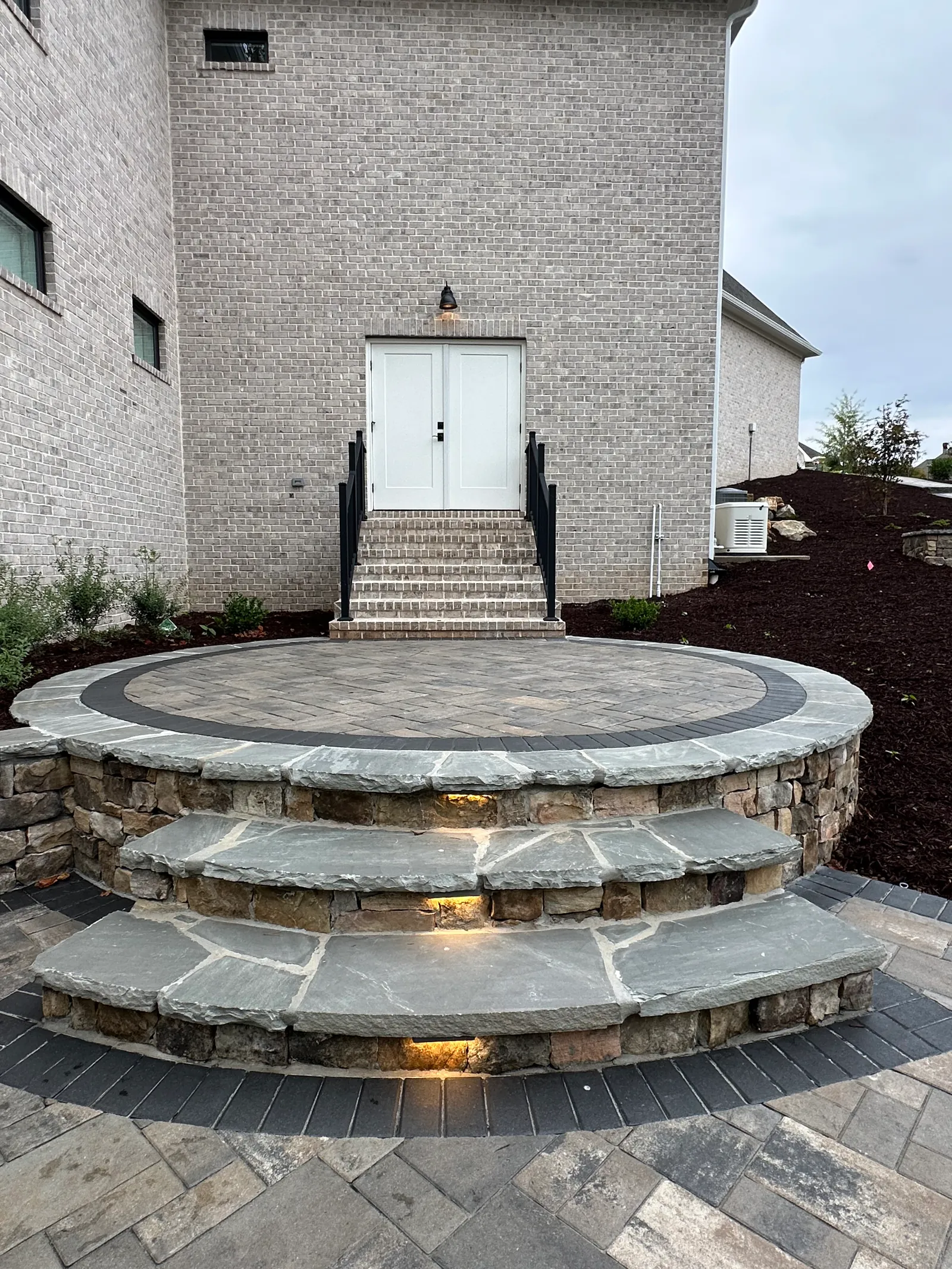 Circular paver patio landing with illuminated curved stone steps leading to a brick home at dusk