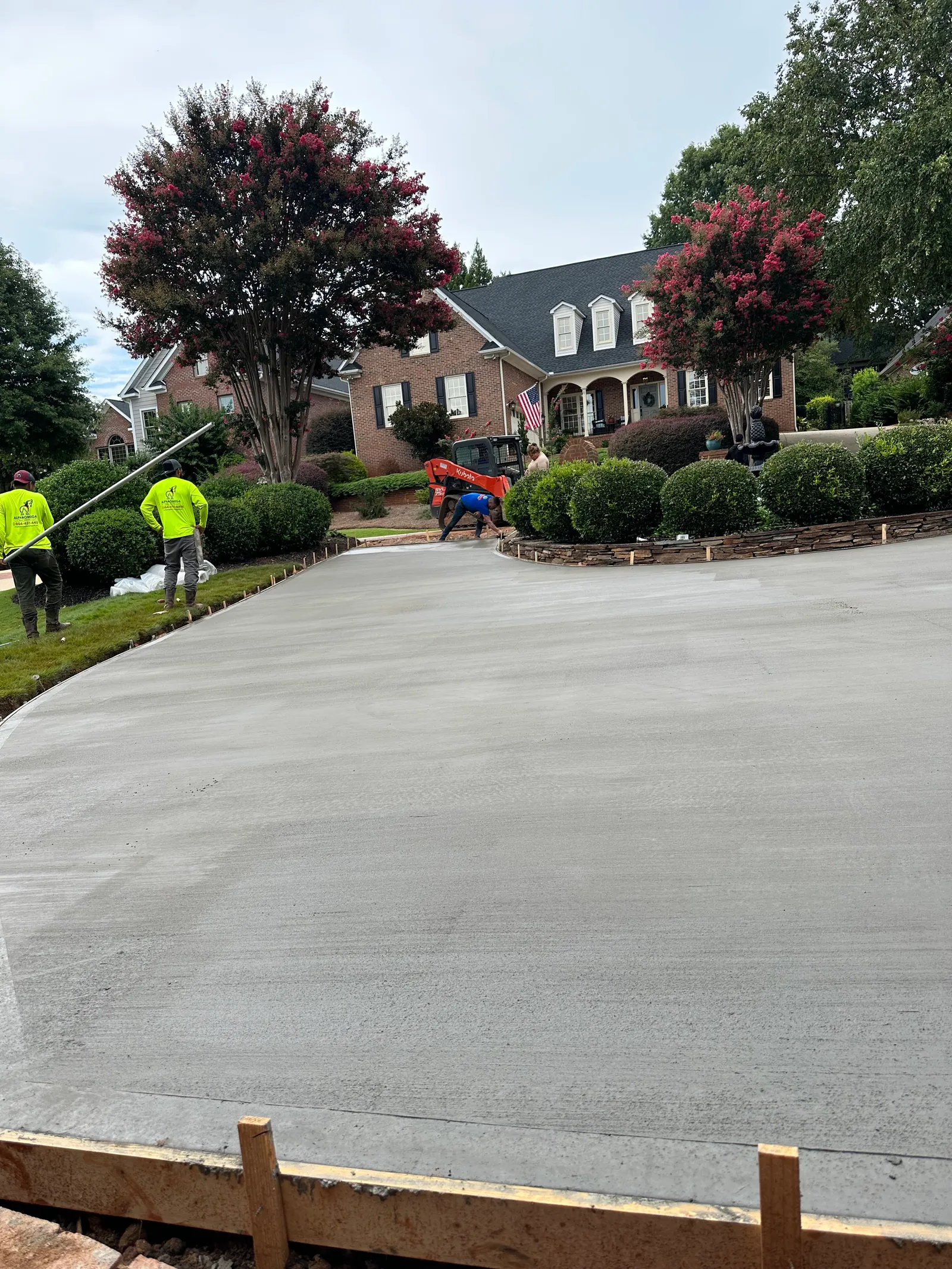 Poured concrete driveway pour in progress, two-story brick home with dormers, crepe myrtle trees