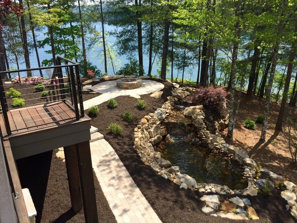 Stacked river-rock waterfall and pond feature next to a stone fire pit, lakefront lot with a cable-rail deck overlooking the water