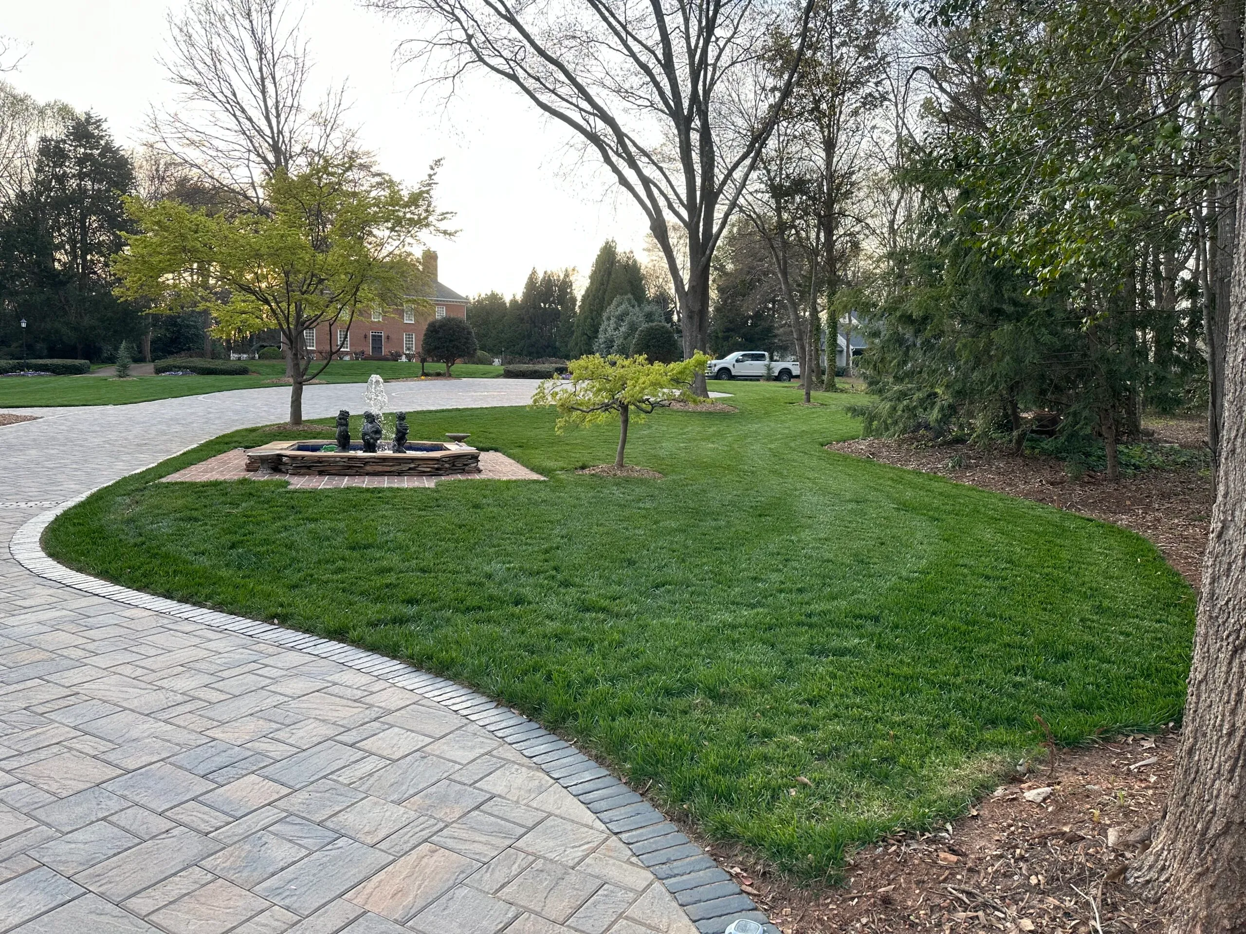 Three-tier stone fountain set in a paver courtyard island, surrounded by manicured lawn