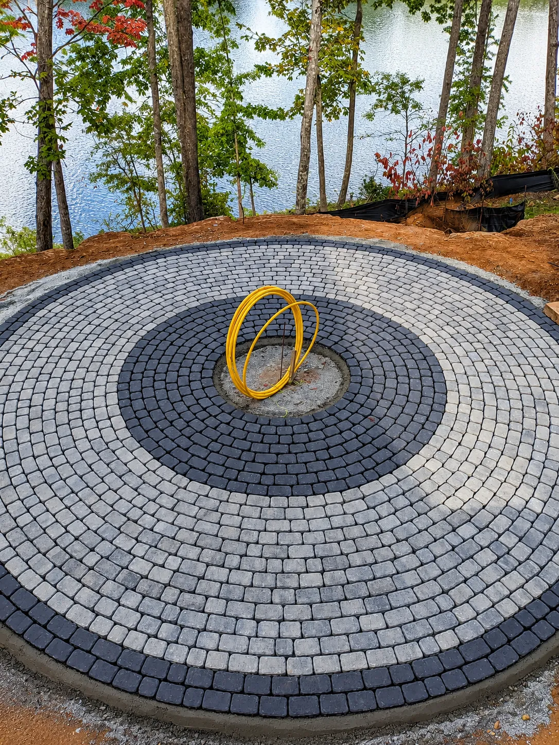 Circular paver patio laid in concentric rings on a wooded lakefront lot, under construction