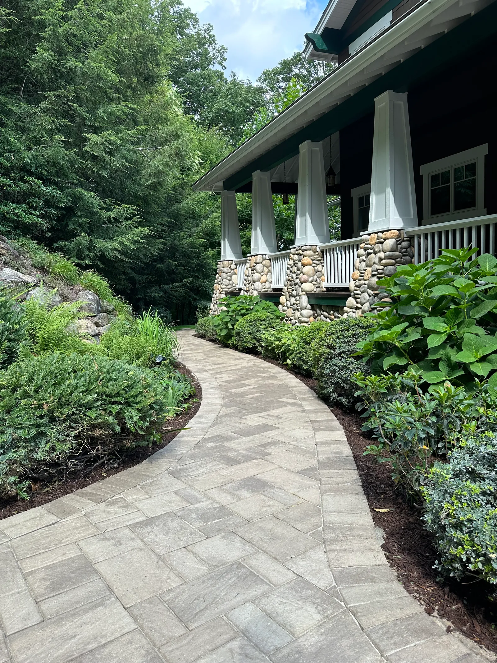 Curving paver walkway through lush planting leading to a craftsman-style home with river-rock porch columns