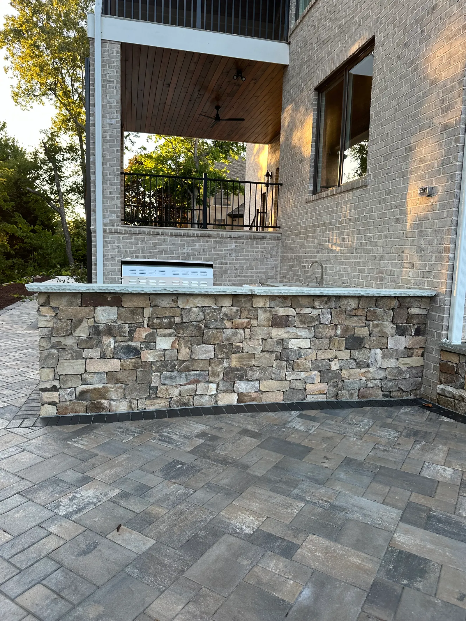 Stone outdoor kitchen counter under construction with a grill still under cover, brick home with a balcony above