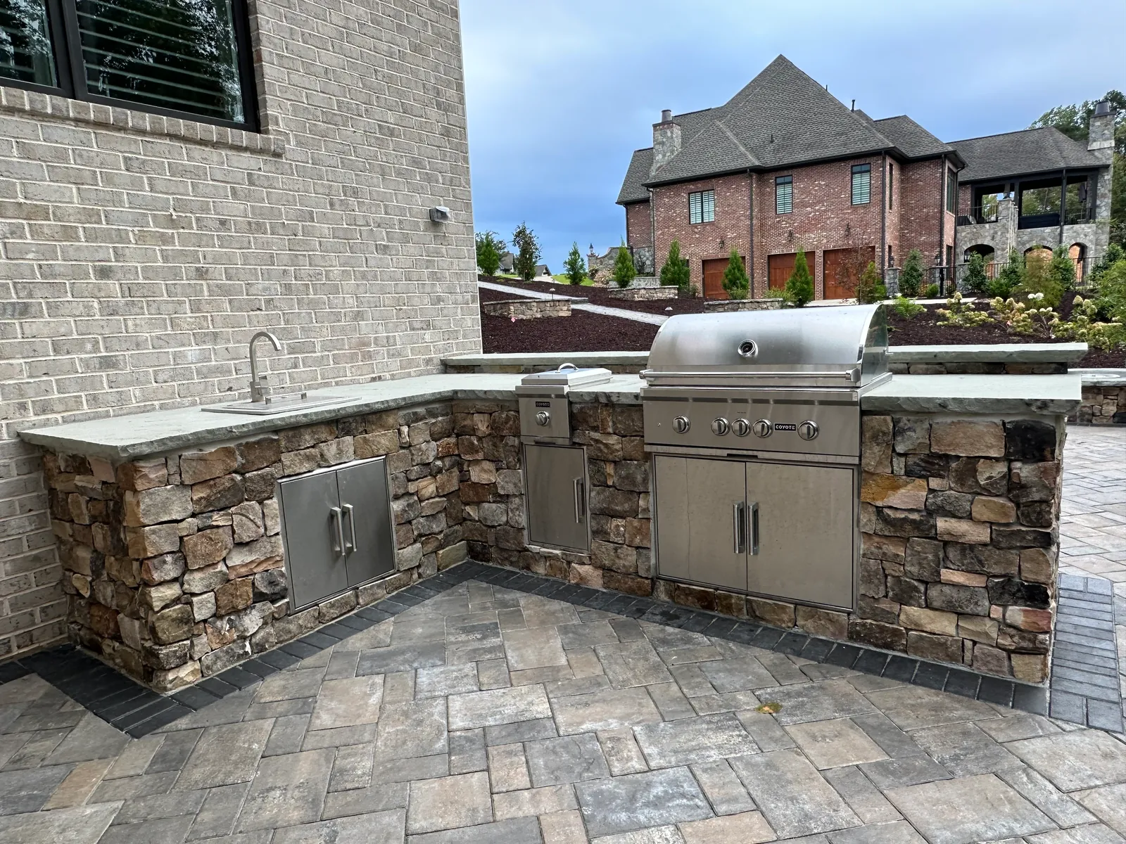 Full stone outdoor kitchen with sink, side burner, and grill, brick estate home visible across a landscaped slope