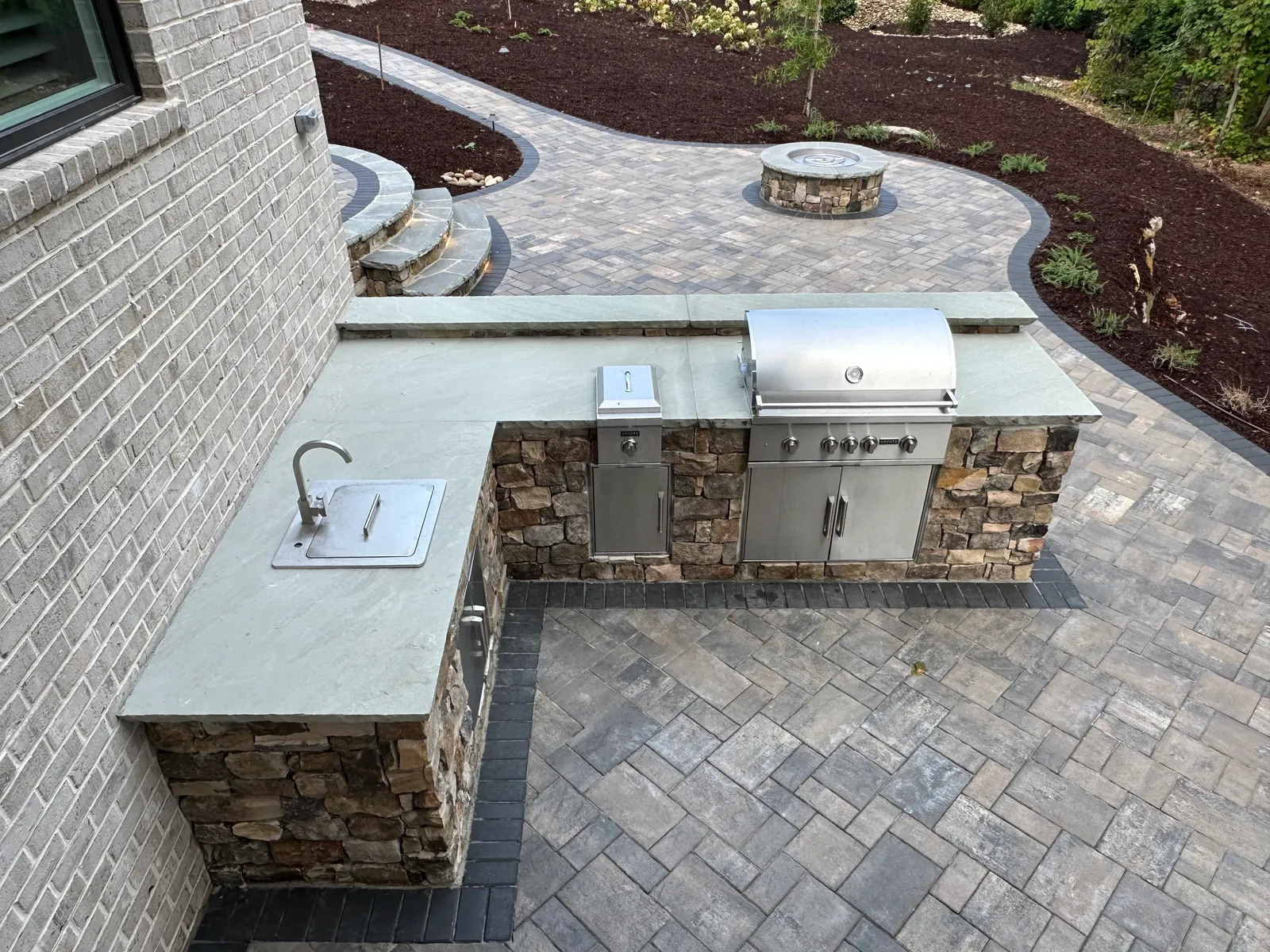 Aerial view of a stone outdoor kitchen connected by a curving paver path to a round fire pit