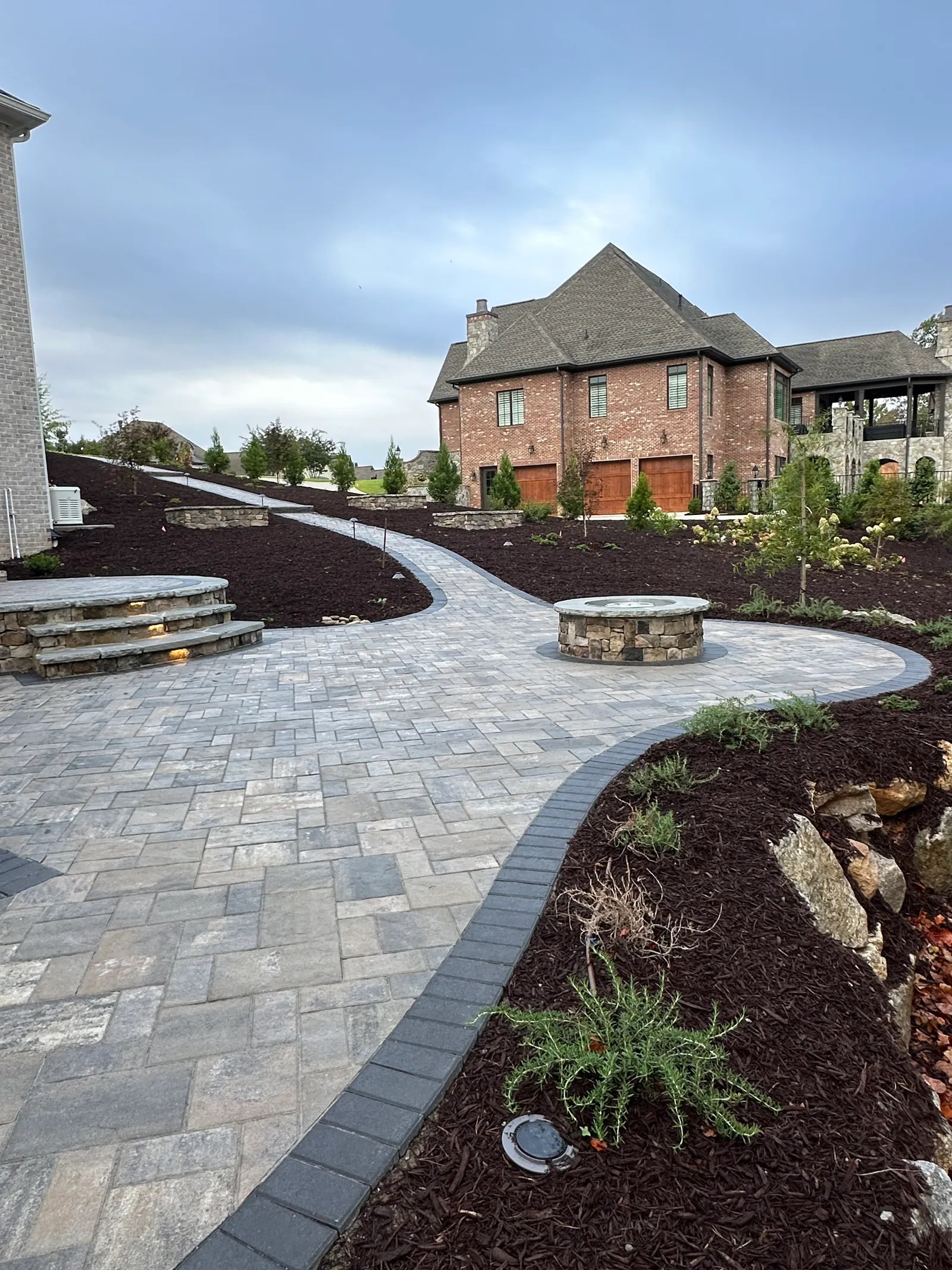 Curving paver walkway past a round stone fire pit toward a large brick estate home with a three-car garage