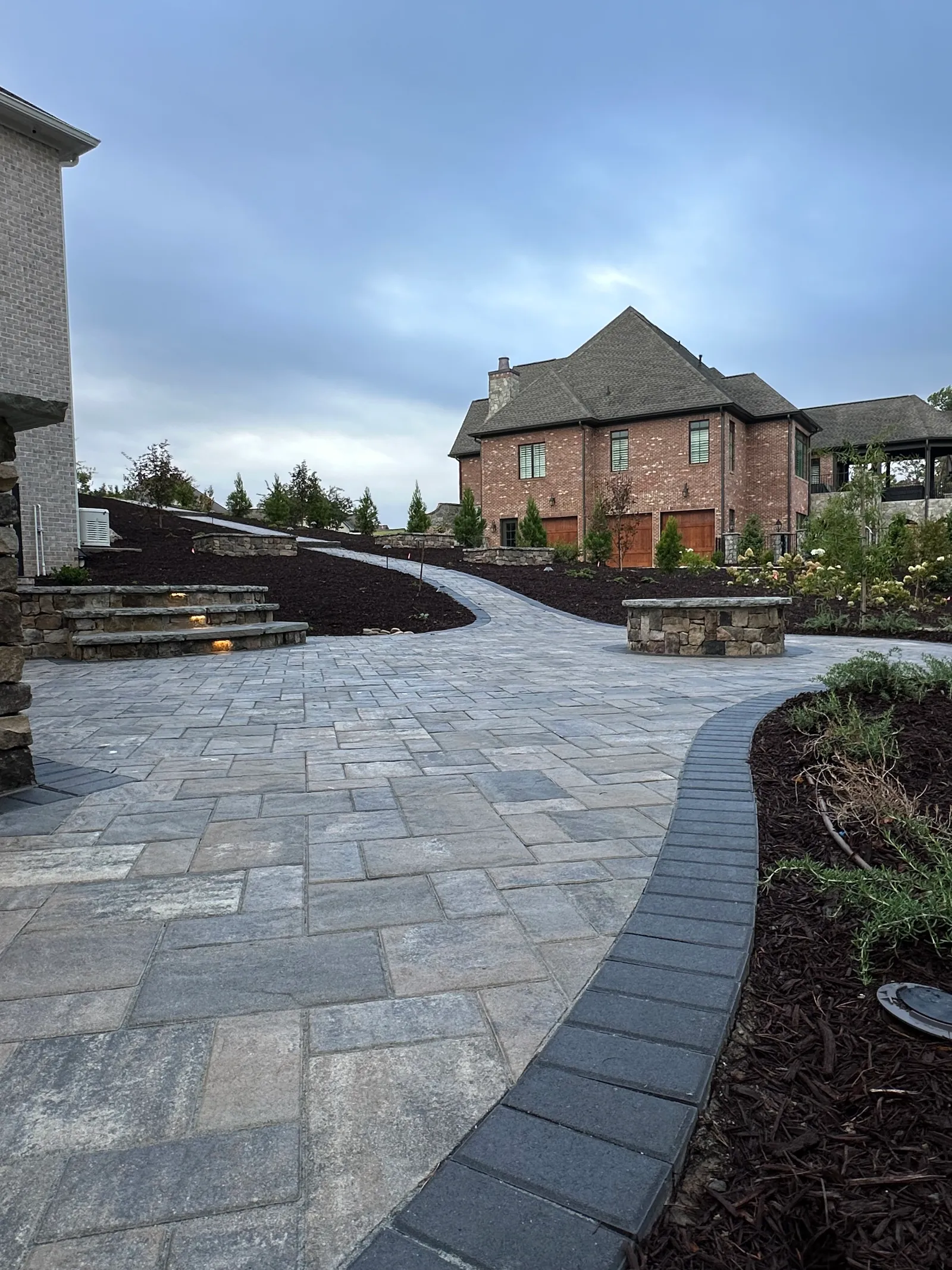 Paver patio with illuminated stone steps and a round fire pit at dusk, brick estate home in the background