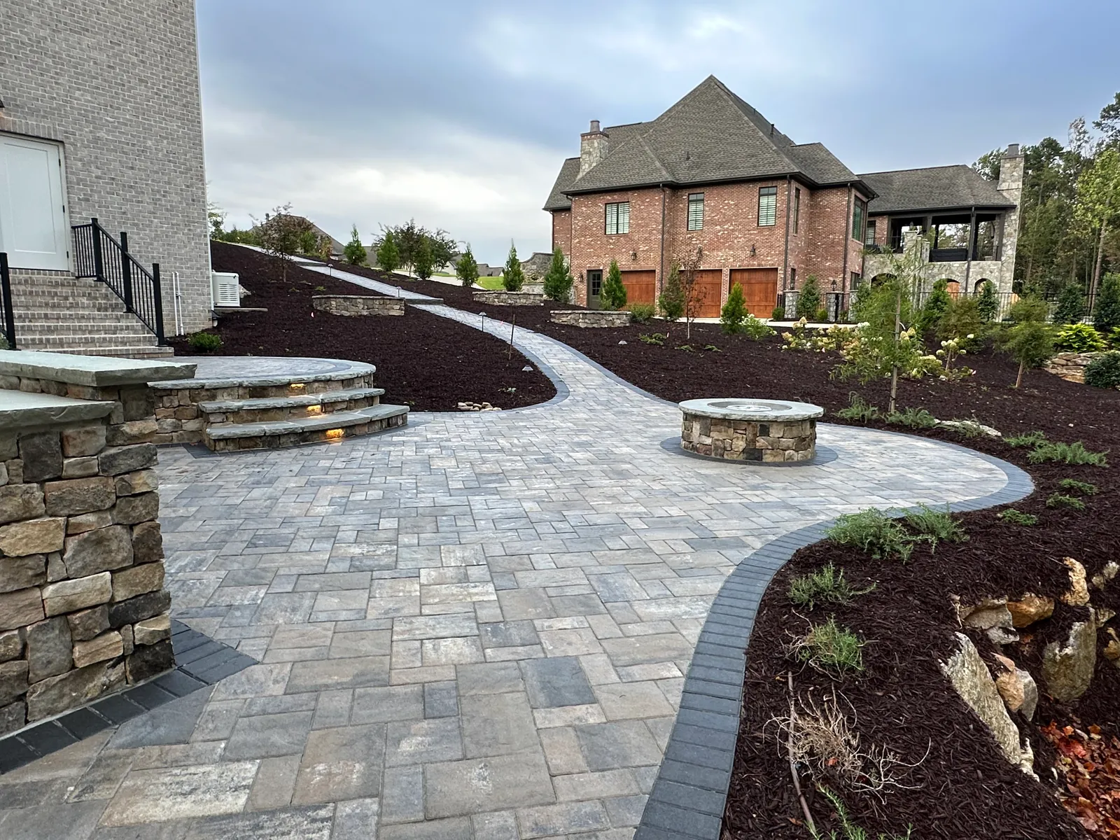 Wide view of a paver patio, stone steps, and fire pit in front of a large brick estate home with a stone pavilion
