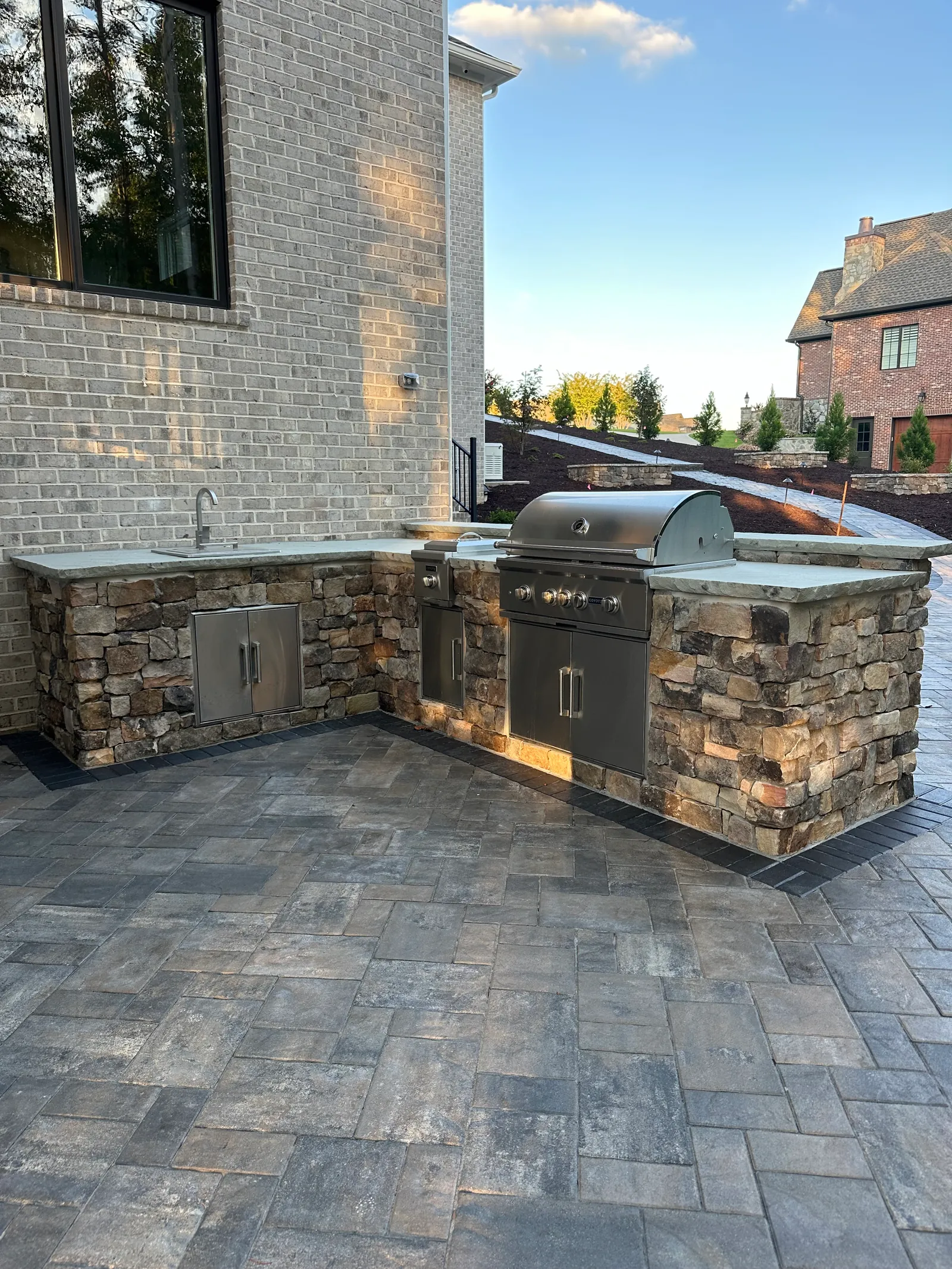 Stone-clad outdoor kitchen with a built-in grill on a paver patio, golden-hour light, estate home in the background