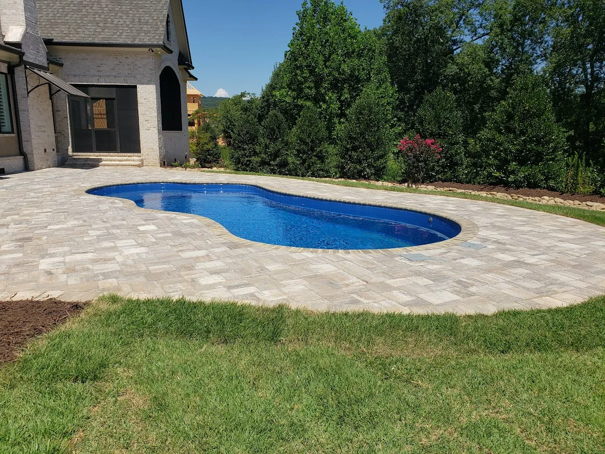 Freeform kidney-shaped pool with a natural stone paver deck, light gray brick home, distant ridgeline visible