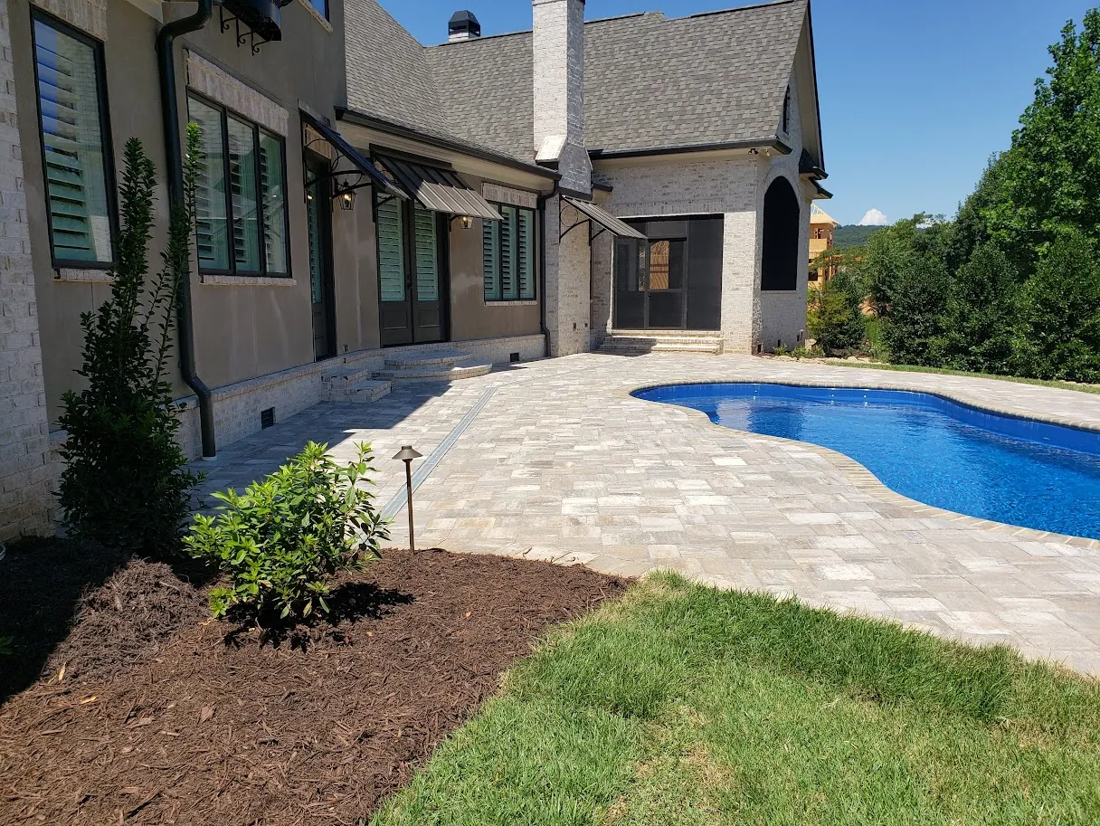 Freeform kidney-shaped pool with a paver deck beside a cream stucco home with dark metal awnings