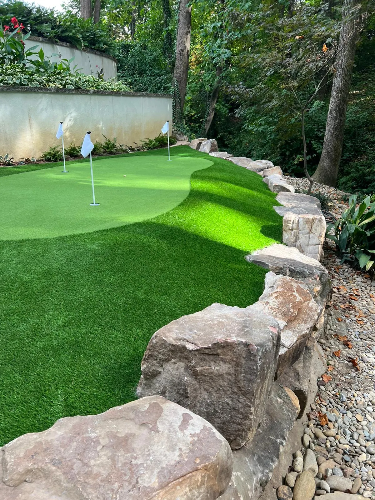 Three-hole artificial turf putting green bordered by large natural boulders on a wooded slope