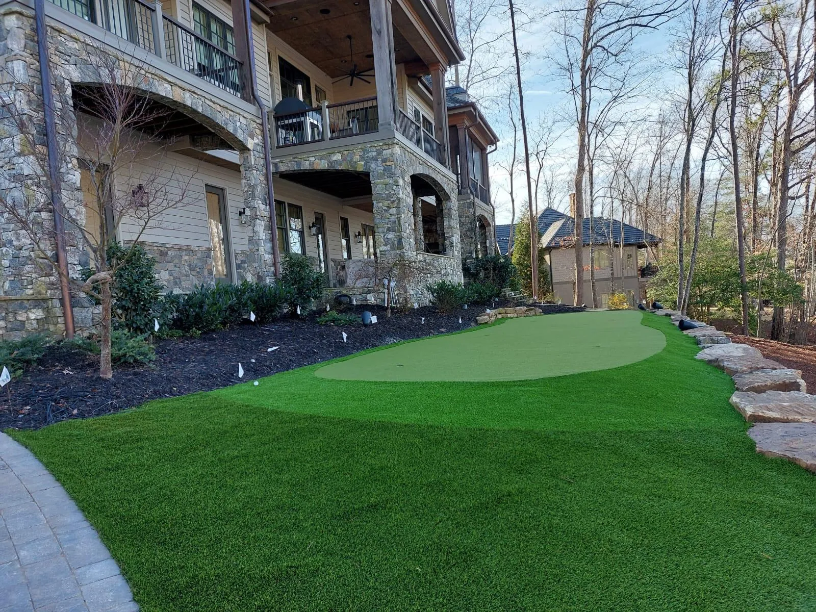 Two-tier artificial turf putting green beside a large stone estate home with a natural stone retaining wall