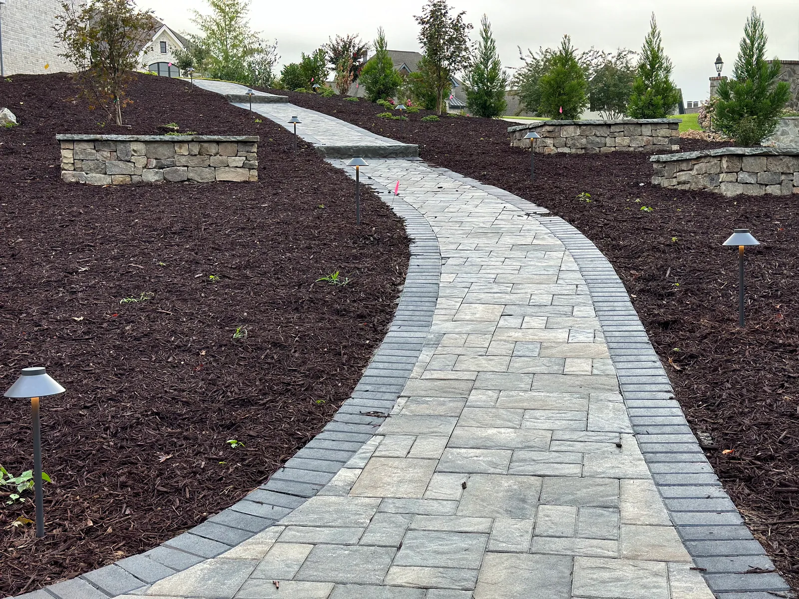 Wide view of a curving paver walkway with matching stone piers and lighting fixtures through fresh planting beds