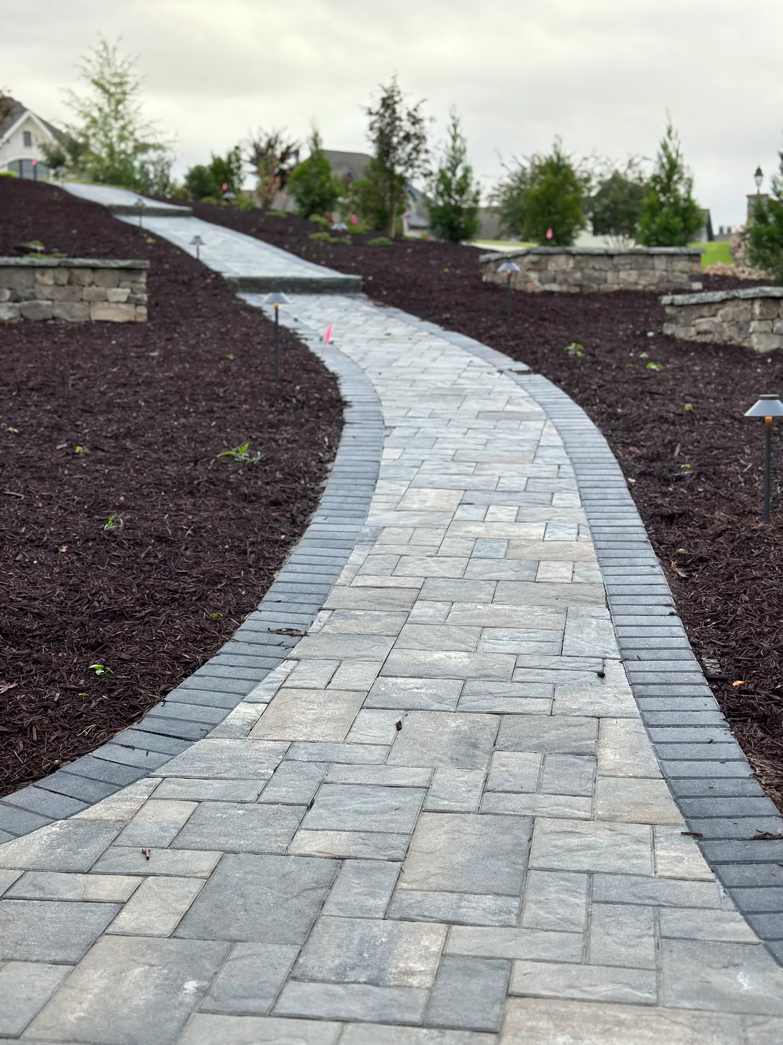 Winding paver walkway with stone-capped piers and landscape lighting curving across a sloped lot