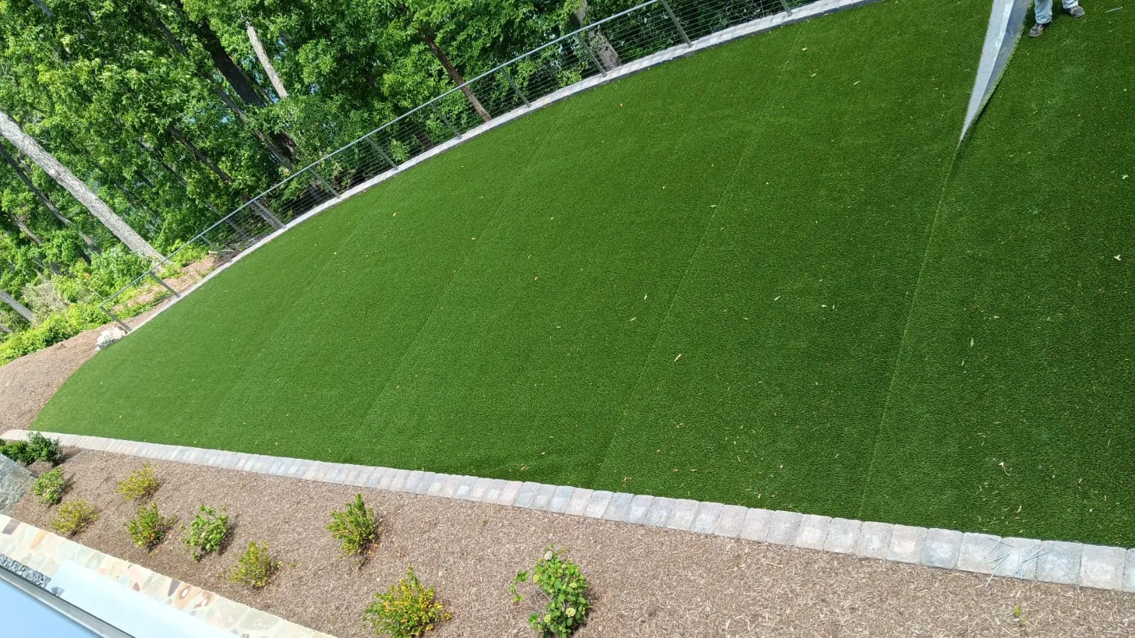 Wide artificial turf lawn on a terraced slope bordered by a cable-rail fence and paver edge