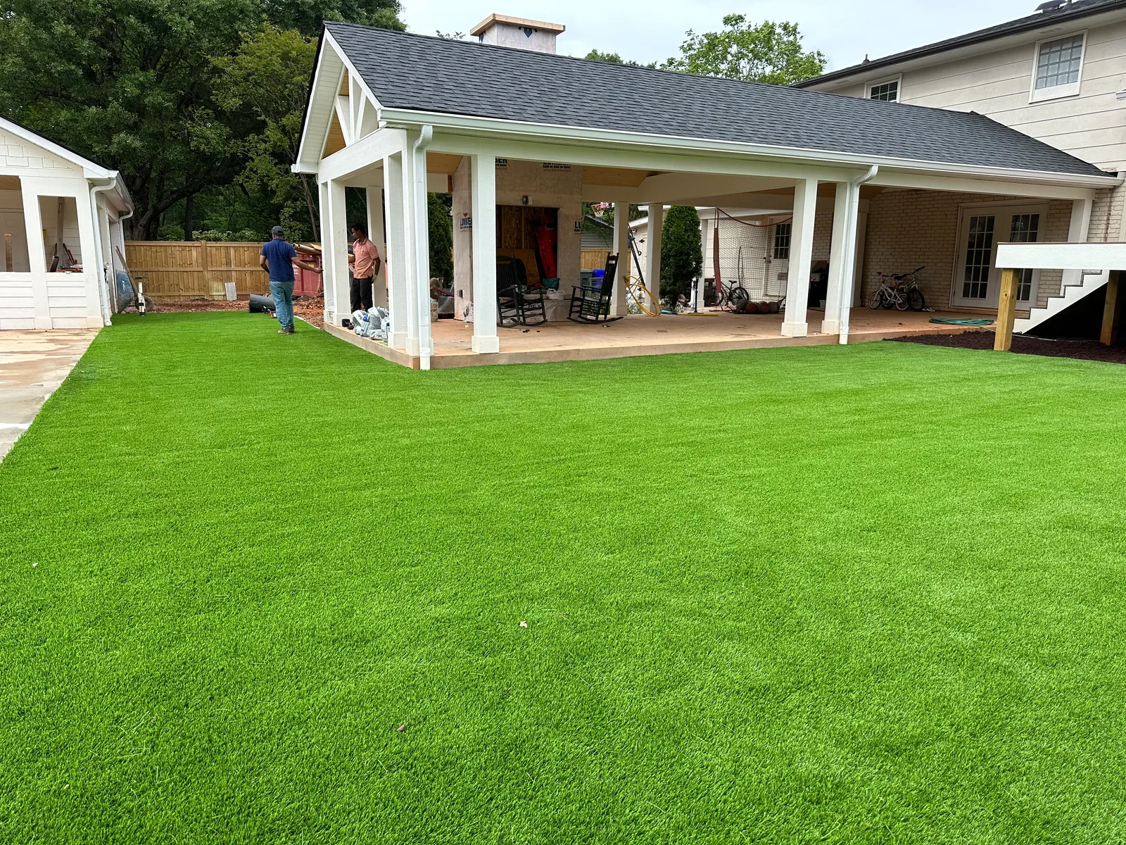 Wide artificial turf lawn beside a covered outdoor pavilion, installation crew finishing the seams